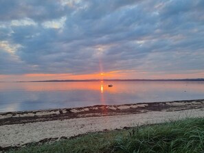 A calm body of water with a sunset, a small boat, and a sandy beach with grass in the foreground. - 4 Star Holiday Home in Ebeltoft (Ebeltoft)