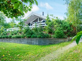A modern house with a sloped roof, surrounded by a well-maintained garden with various shrubs and a stone retaining wall.