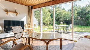 Sala de estar moderna com teto de madeira, uma televisão grande, uma mesa redonda e vista para um jardim através de amplas janelas.