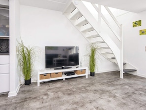 A modern living room with a flat-screen TV on a white stand, a staircase, and two potted plants.