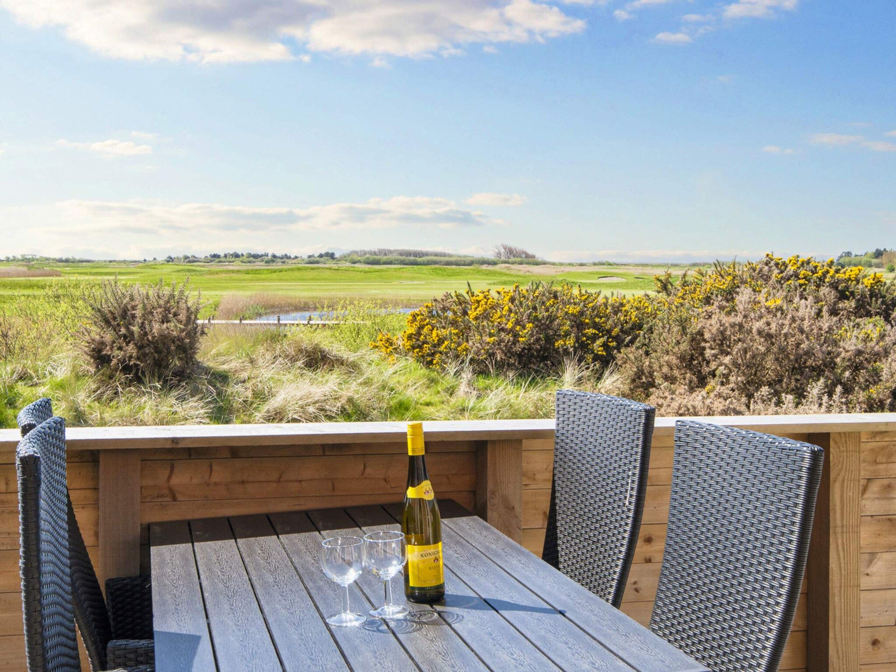 A wooden table with two chairs and a bottle of wine, overlooking a grassy landscape with a clear sky.