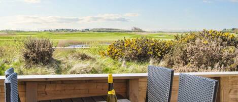 A wooden table with two chairs and a bottle of wine, overlooking a grassy landscape with a clear sky.