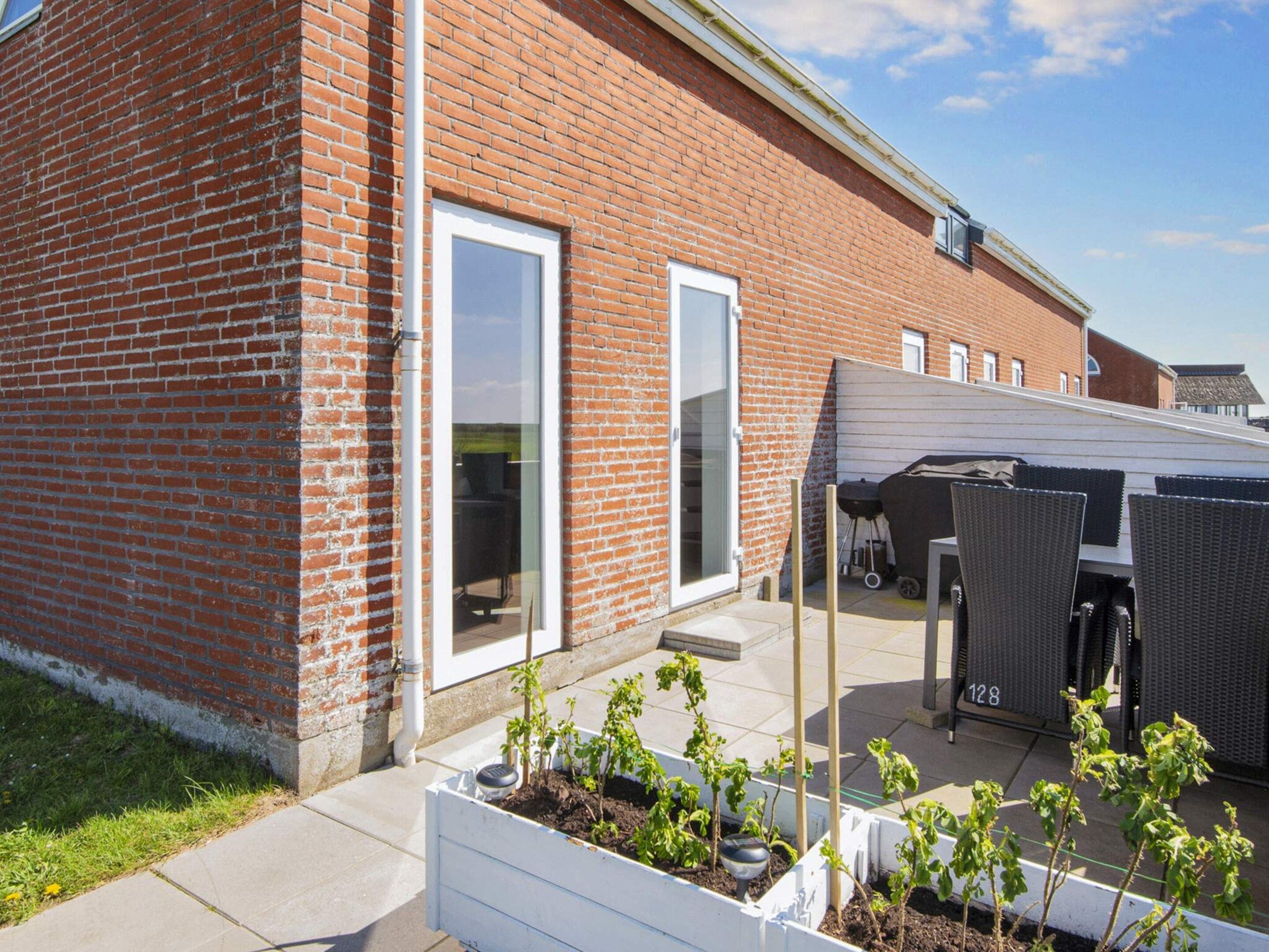 A brick building with a white door and windows, a covered outdoor seating area, and raised garden beds with plants.
