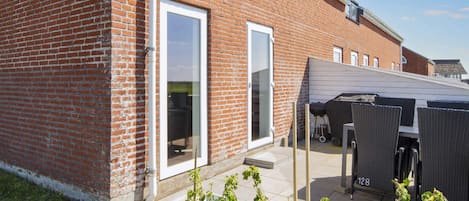 A brick building with a white door and windows, a covered outdoor seating area, and raised garden beds with plants.