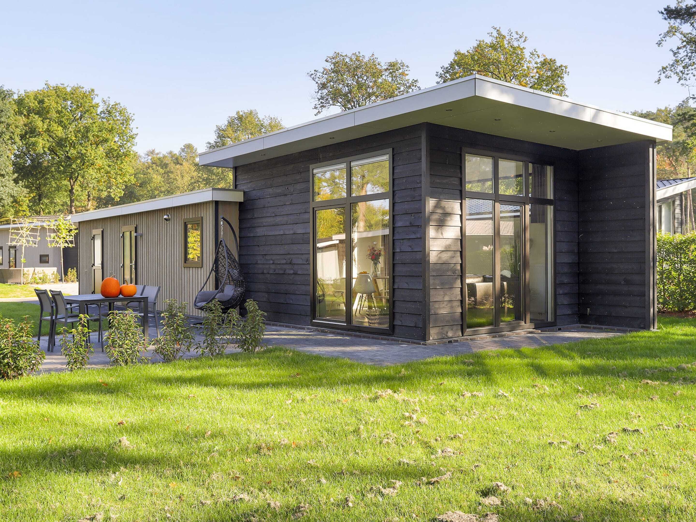 Une maison moderne dotée d’une grande baie vitrée, d’un patio avec une table et des chaises, et d’un fauteuil suspendu.