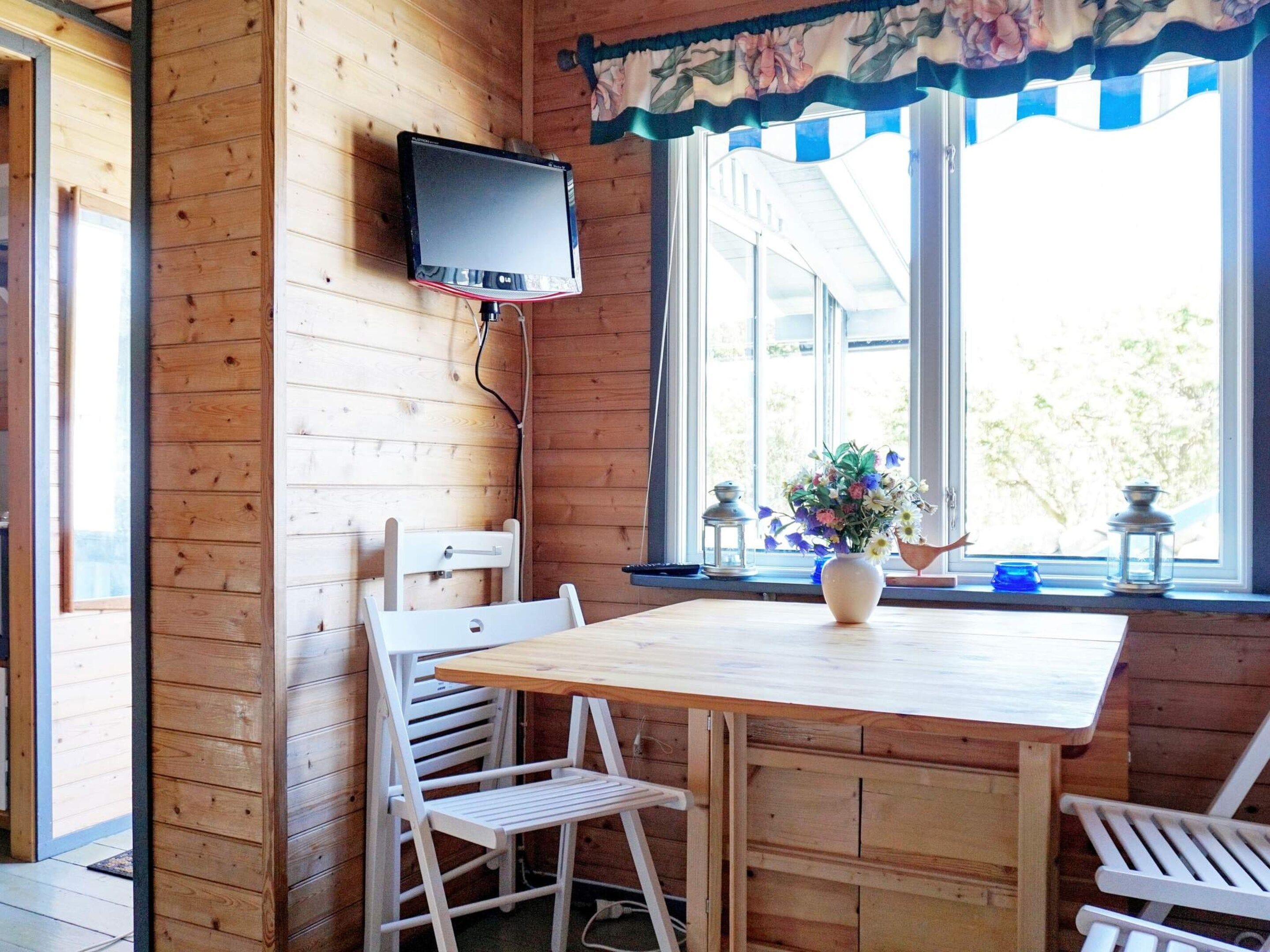 A wooden interior with a TV mounted on the wall, a table with chairs, and a window with a view of greenery.