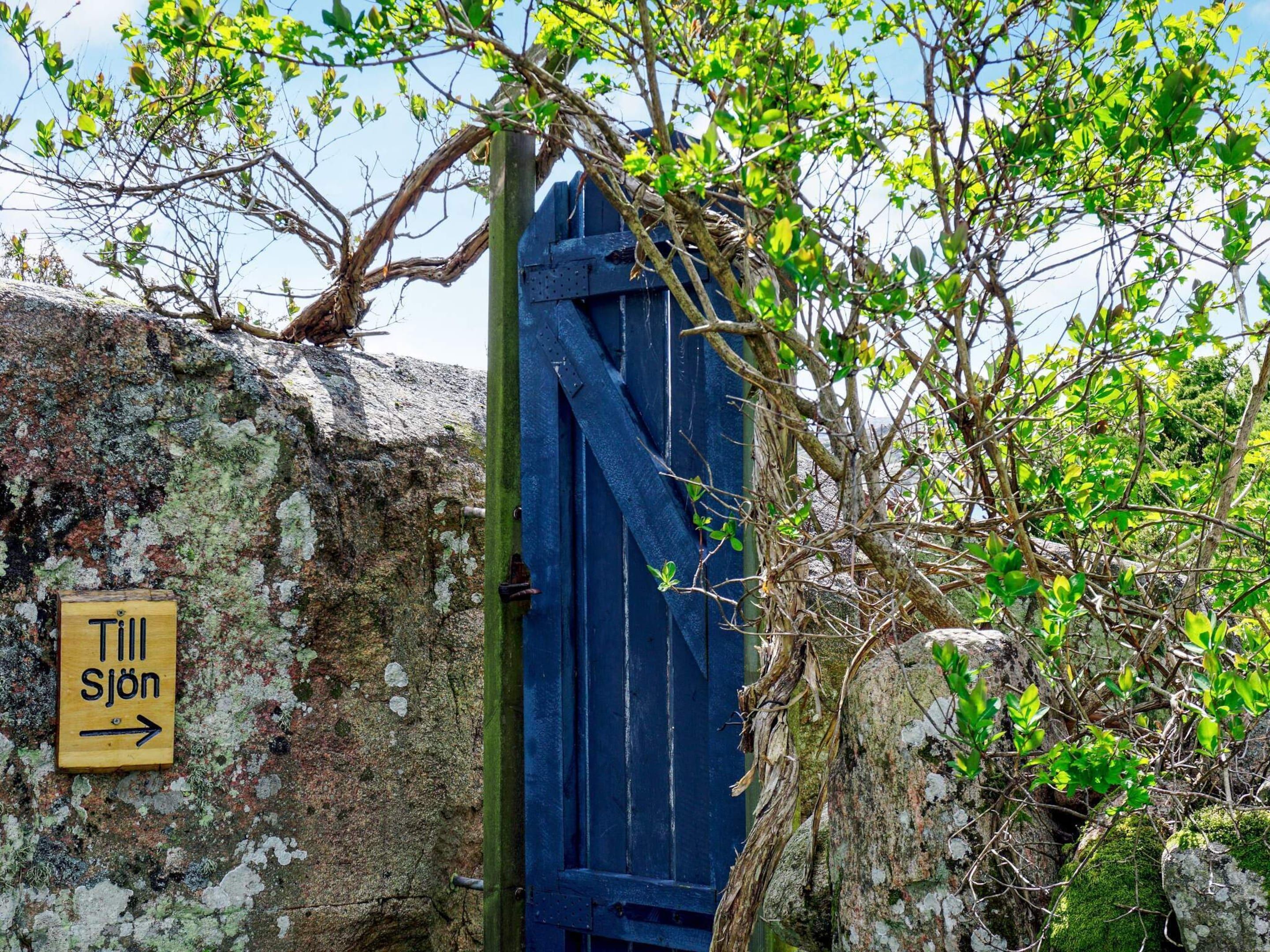 A rustic wooden door with a sign reading "Till Sjön" pointing to the right, surrounded by moss-covered rocks and green foliage.