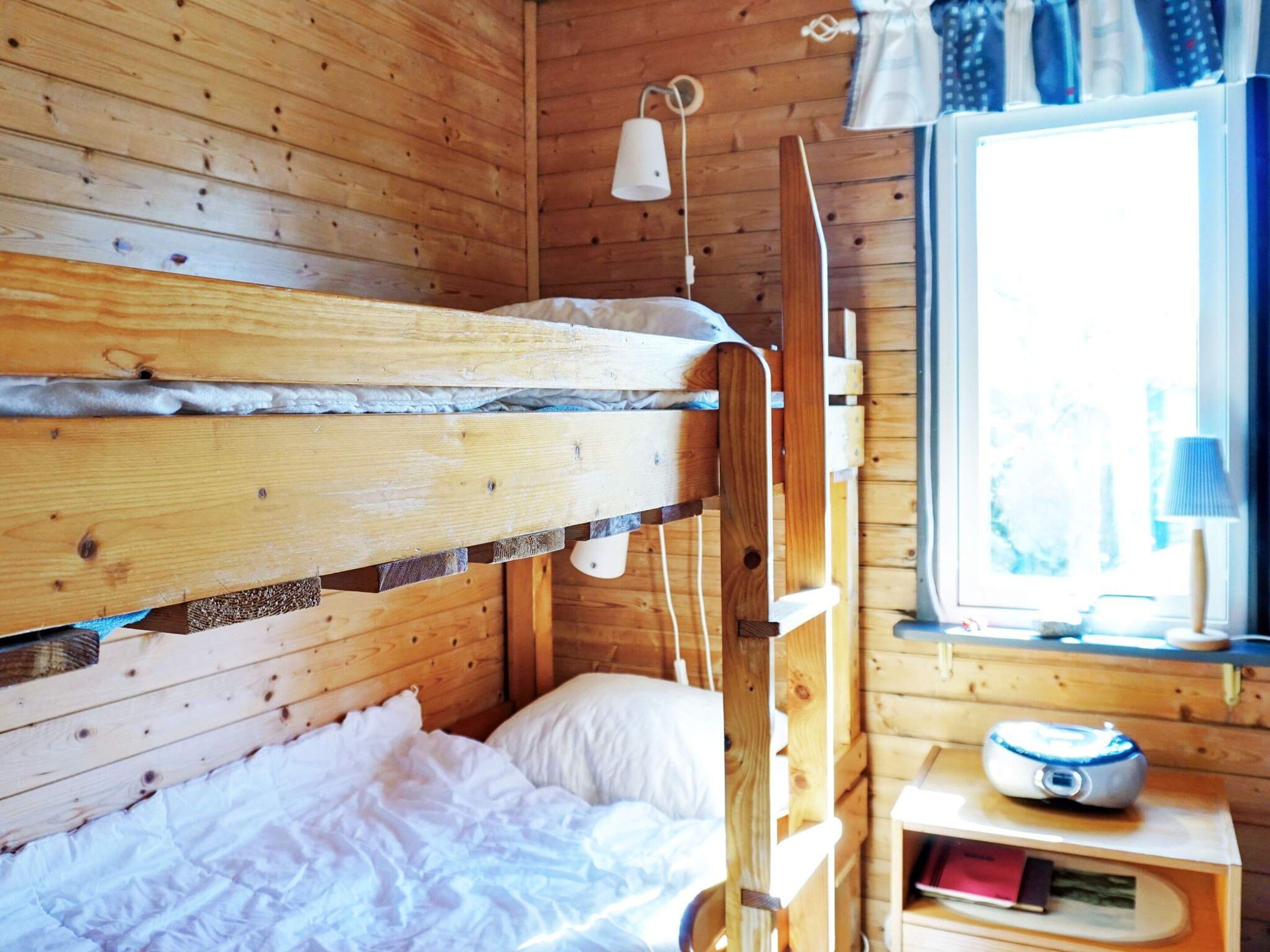 A wooden bunk bed with a white mattress and pillow, a bedside table with a lamp, and a window with curtains.