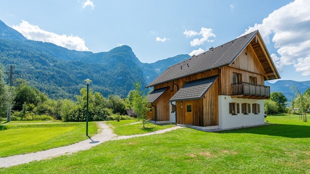 Uma casa de madeira de dois andares com varanda, rodeada por uma paisagem verde e montanhas ao fundo.