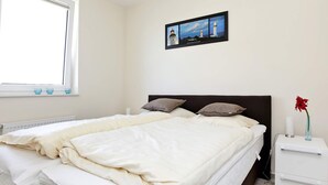 Un dormitorio con cama, almohadas, una mesita de noche, un florero con una flor y un cuadro en la pared.