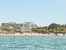 A coastal hotel with multiple floors, a beach in the foreground, and a bridge in the background.