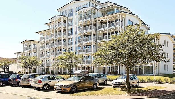 Un edificio residencial de varios pisos con balcones, coches aparcados y árboles.