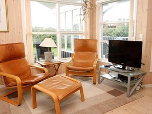 A living room with two leather chairs, a small table, a television on a stand, and a view of the outdoors through large windows.