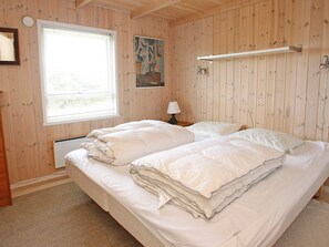 A room with wooden walls, a bed with white bedding, a window with blinds, a painting on the wall, and a bedside lamp.