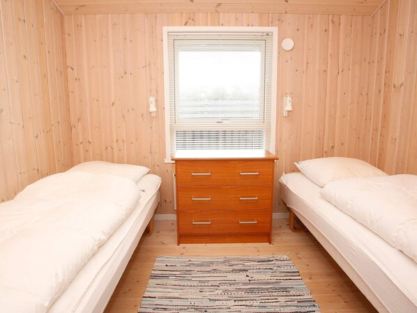 A room with two single beds, a wooden dresser with drawers, a window with blinds, and a patterned rug on the floor.