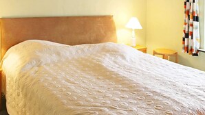 A neatly made bed with a textured white duvet, a wooden headboard, a bedside lamp, and a small wooden stool. - Cozy Holiday Home in Nexo near Dueodde Beach (Nexø)