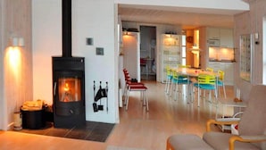 A cozy living room with a fireplace, a dining area with colorful chairs, and a kitchen in the background. - 8 Person Holiday Home in Lokken (Lokken)