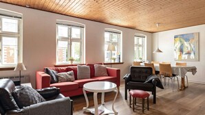 Een ruime woonkamer met een rode bank, een ronde witte tafel en een zwarte leren fauteuil. Er zijn meerdere ramen die zorgen voor natuurlijk licht, en een schilderij hangt aan de muur.