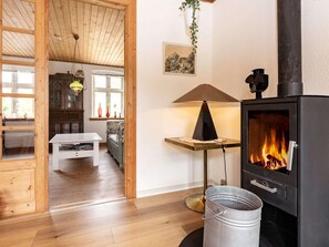 A cozy living room with a wooden floor, a white table, a sofa, a black lamp, a fireplace with a visible fire, and a hanging plant. - 6 Person Holiday Home in Marstal (Marstal)
