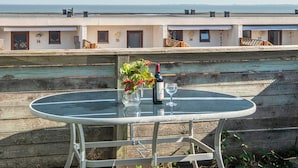 A round glass table with a bottle and glass on it, set against a wooden deck with a view of a building with numbered windows.