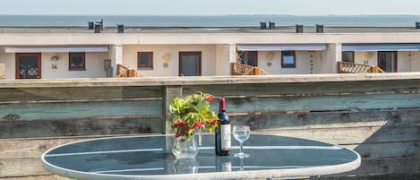 A round glass table with a bottle and glass on it, set on a wooden deck overlooking a waterfront with buildings in the background.