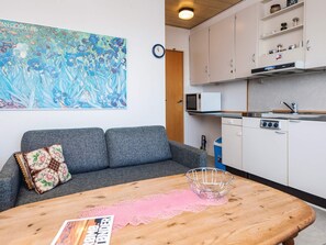 A room with a grey sofa, a wooden table with a pink doily, a metal bowl, and a kitchen area with white cabinets and a microwave.