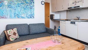 A compact living space with a grey sofa, a wooden table with a pink doily, a metal bowl, and a kitchen area with white cabinets, a microwave, and a clock.