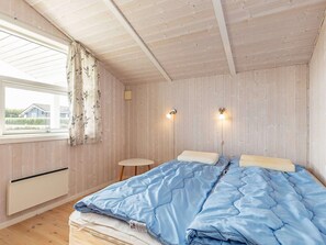 Et soveværelse med træloft, en seng med blåt sengetøj, to sengeborde med lamper, et vindue med gardiner og et lille natbord.