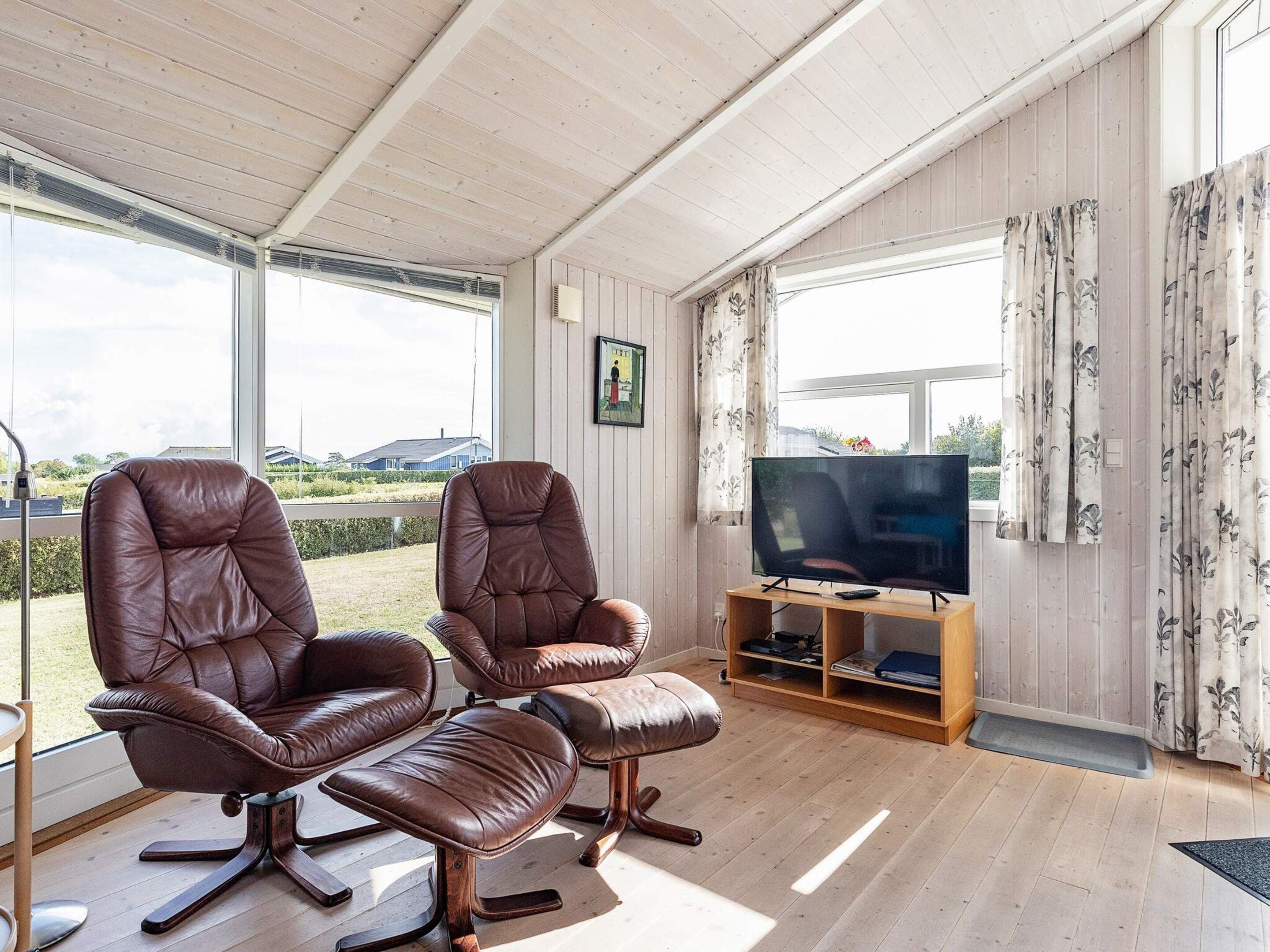 A spacious living area with two leather armchairs, a television on a wooden stand, and large windows offering a view of the outdoors.