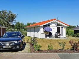 A small, single-story house with a red-tiled roof, a covered patio with outdoor furniture, and a parked car in the driveway.