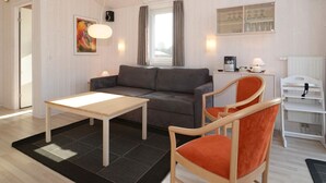 A modern living room with a sofa, a table, and a chair. There is a window with curtains, a radiator, and a shelf with glasses. - Holiday Home in GrÃ¶mmitz (Groemitz)