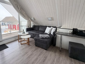 A modern loft-style room with a grey sofa, a small round table, and a kitchenette area. - Holiday Home in Wendtorf (Wendtorf)