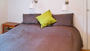 A bed with a grey quilt, two grey pillows, and a green pillow. There are two wall-mounted lamps. A small wooden nightstand with a book is beside the bed. - 4 Star Holiday Home in Sturefors (Sturefors)