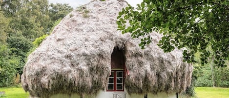 Casa con techo de paja, una ventana y una puerta a la vista, rodeada de césped y árboles.