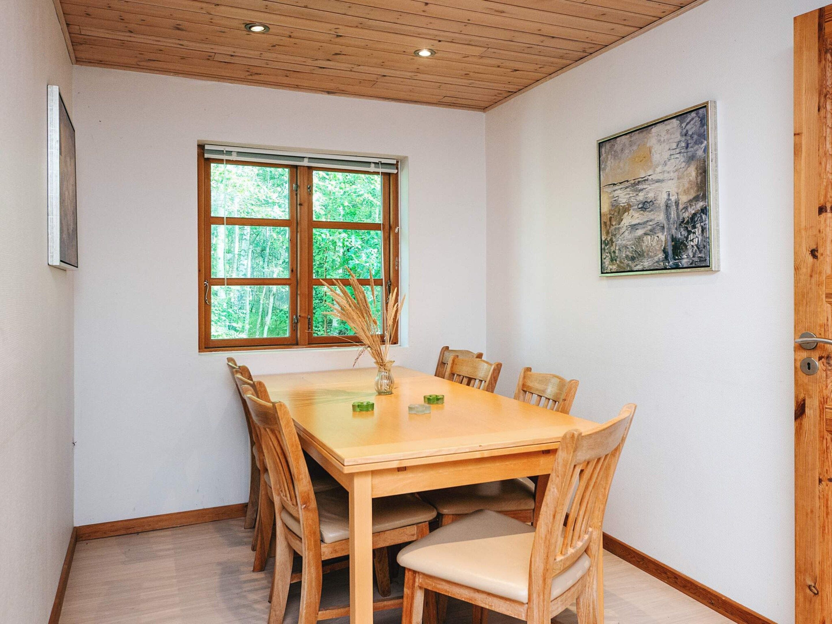 Un coin repas avec une table et des chaises en bois, une fenêtre donnant sur un paysage verdoyant et un tableau accroché au mur.
