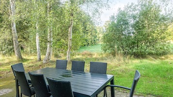 Un área de comedor al aire libre con mesa y sillas, rodeada de árboles y un campo cubierto de césped.