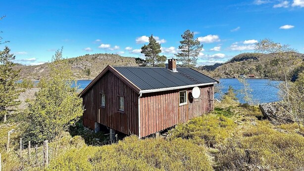 A wooden cabin with a metal roof, situated by a lake with hills in the background. - 5 Person Holiday Home in Farsund (Farsund)