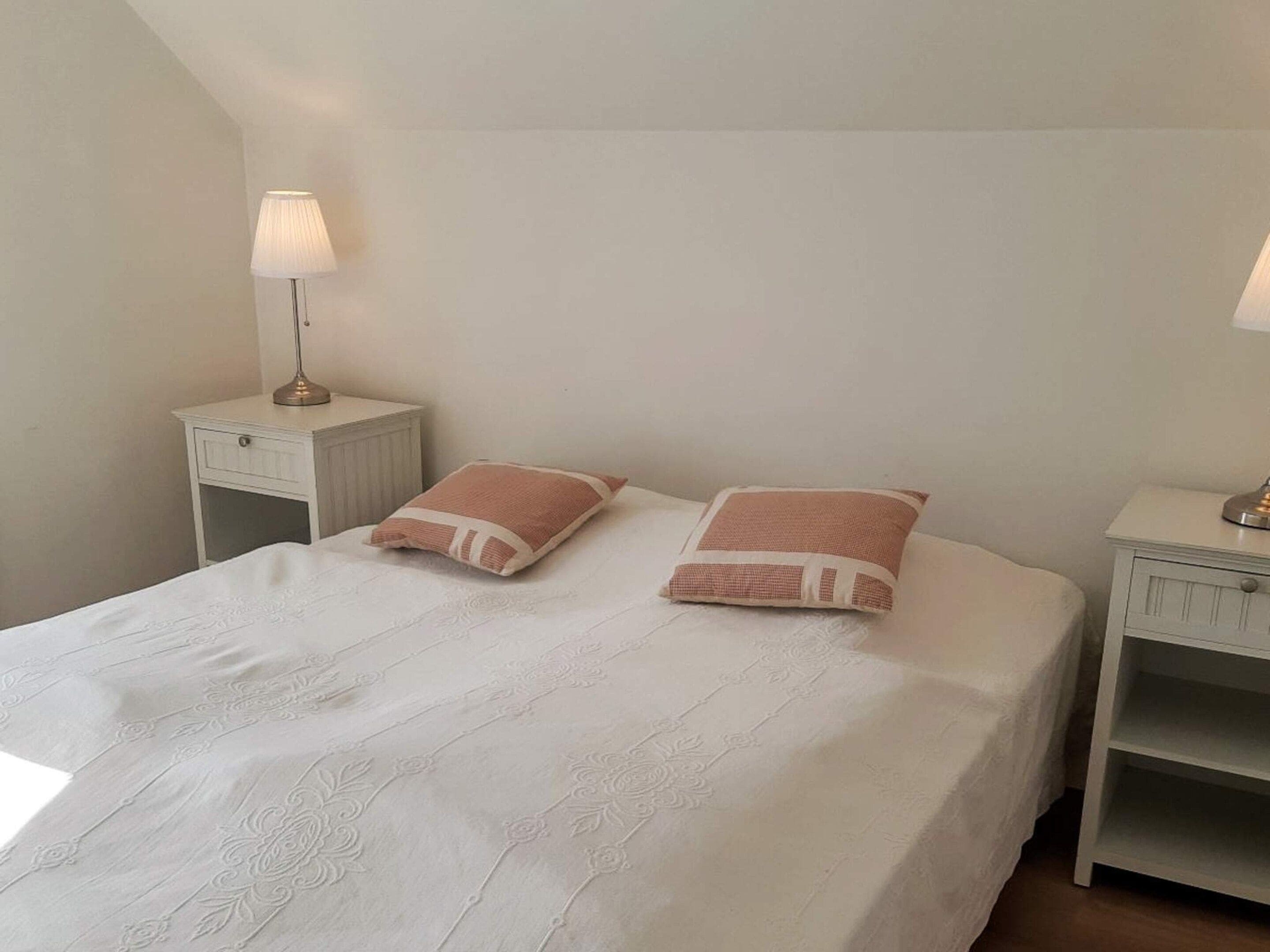 Una cama bien hecha con una colcha blanca y dos almohadas, flanqueada por dos mesitas de noche con lámparas.