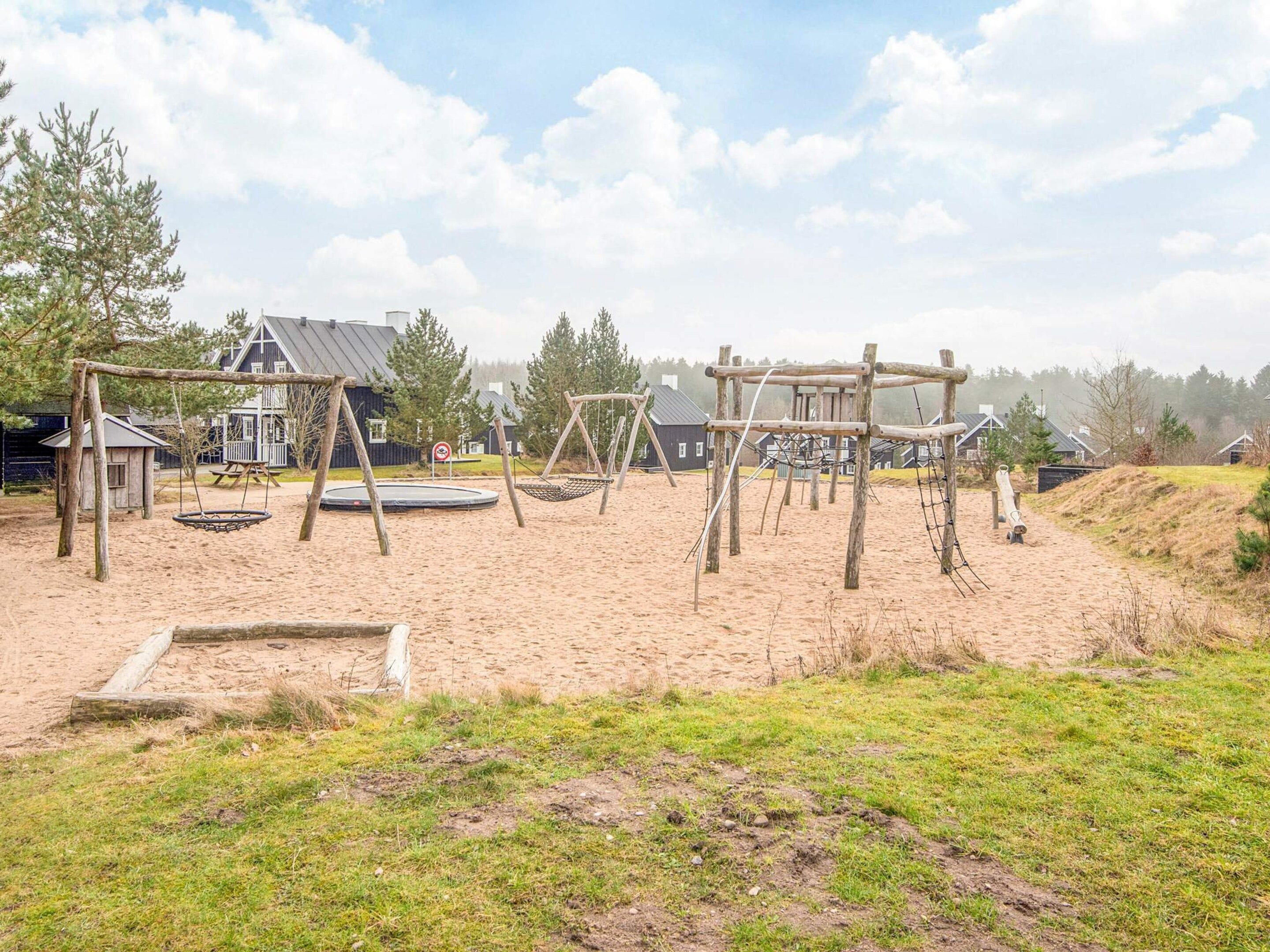 Un parque infantil con columpios y un trampolín, rodeado de arena y césped, con casas al fondo.