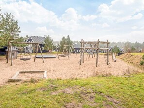 Een speeltuin met schommels en een trampo, omringd door zand en gras, met huizen op de achtergrond.