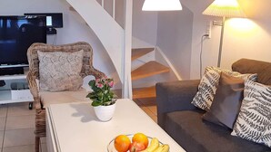 Un saló modern amb un sofà blanc, una cadira de vimetes, una taula de centre de vidre amb fruita, una televisió i una escala.