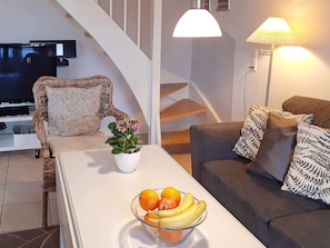 A modern living room with a white sofa, a wicker chair, a glass coffee table with fruit, a TV, and a staircase.