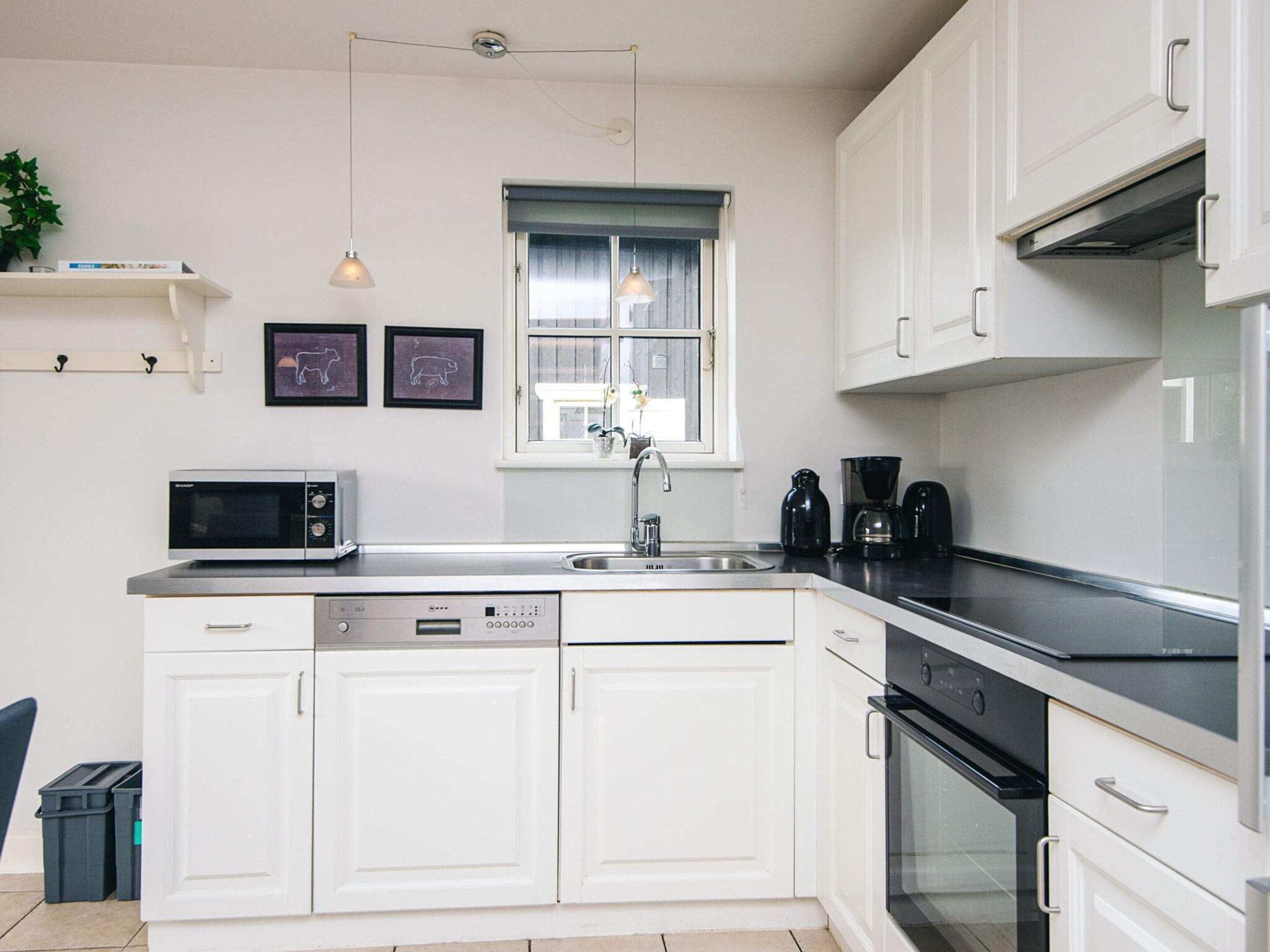 Una cocina moderna con armarios blancos, microondas integrado, lavavajillas, cafetera y una ventana con cortinas.