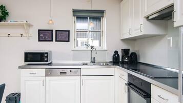 Una cocina moderna con armarios blancos, microondas integrado, lavavajillas, cafetera y una ventana con cortinas.