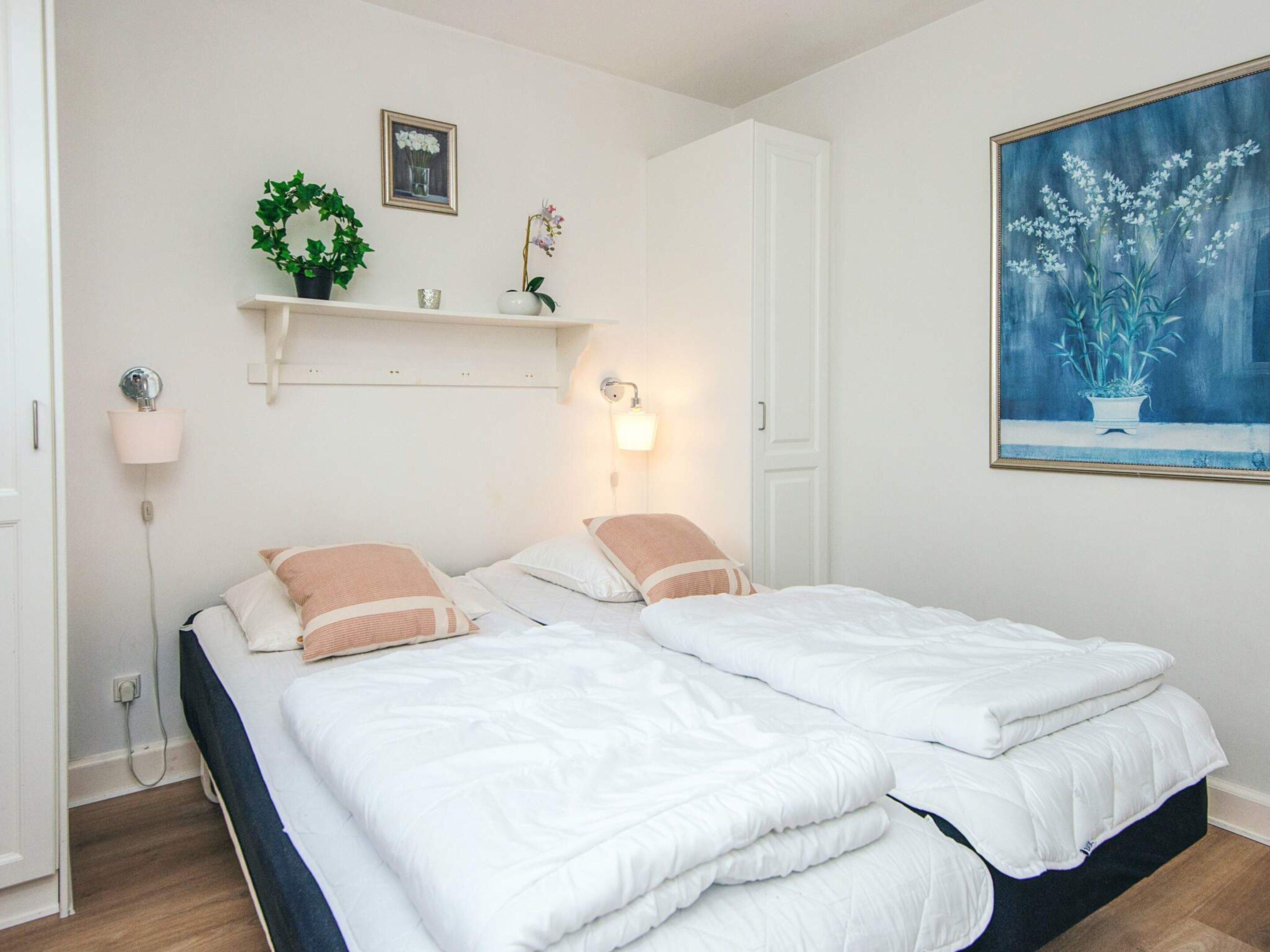 A double bed with white bedding and two pillows, a bedside table with a lamp, a wall-mounted shelf with decorative items, and a framed painting of flowers.