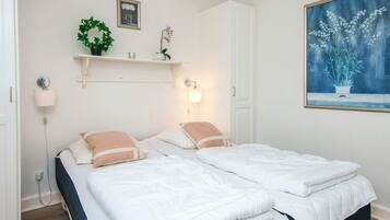A double bed with white bedding and two pillows, a bedside table with a lamp, a wall-mounted shelf with decorative items, and a framed painting of flowers.