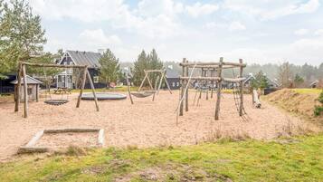 A playground with swings and a trampoline, surrounded by sand and grass.