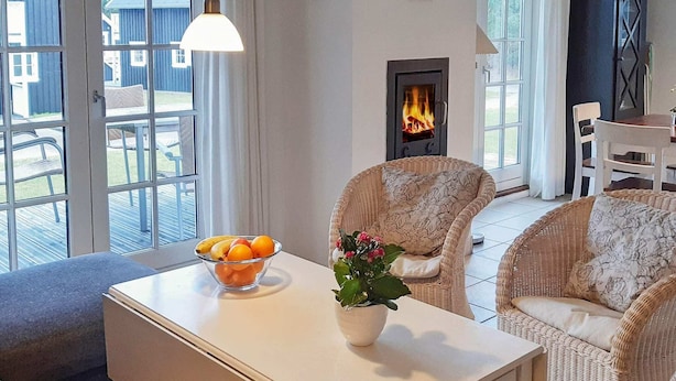 Una sala de estar acogedora con muebles de mimbre, una mesa blanca con un frutero y una planta en maceta, y una chimenea al fondo.