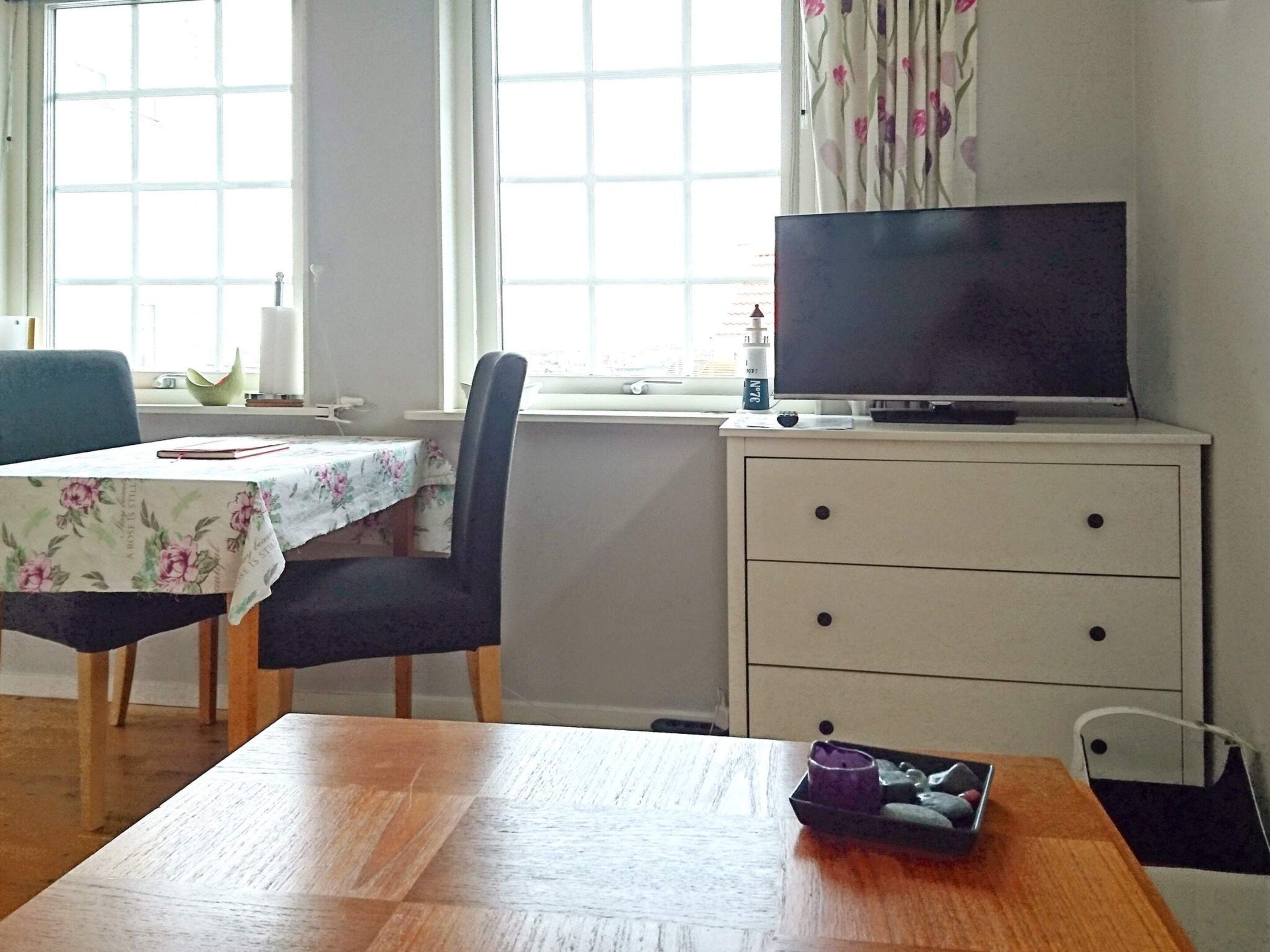 Una habitación con una mesa de madera, una silla, un televisor sobre un mueble y una ventana con cortinas.