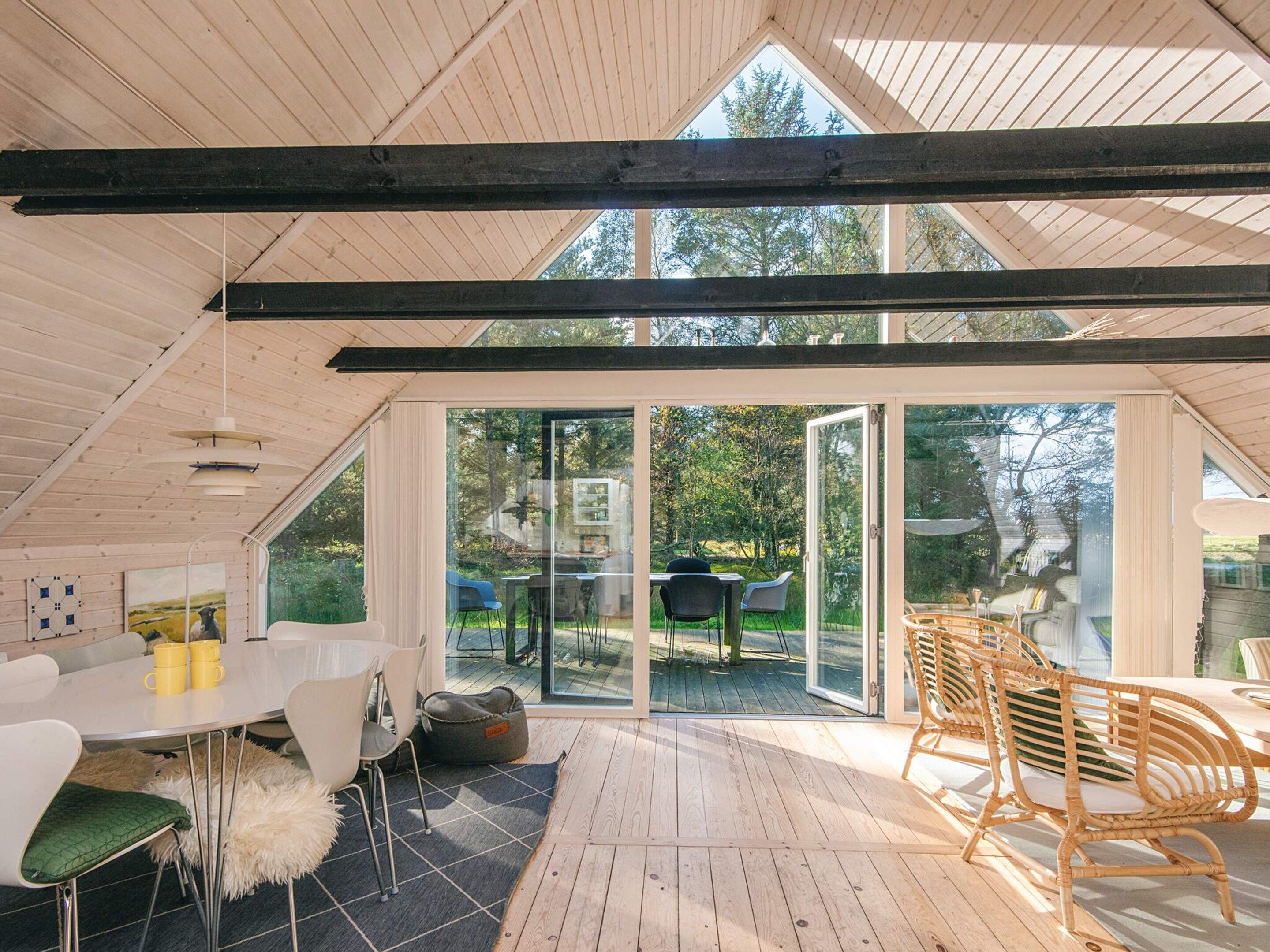 A modern interior with a high ceiling, wooden beams, a dining area with white chairs, a table with yellow cups, and a view of the outdoors through large glass doors.